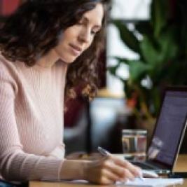 Person sitting in front of a laptop computer writing with a pen and paper.